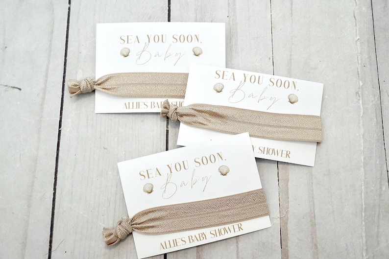 May include: Three white cards with the text "SEA YOU SOON, Baby" and a seashell graphic. Each card has a tan elastic hair tie attached. The text "ALLIE'S BABY SHOWER" is printed on the bottom of one card.