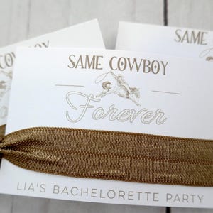 K&ouml;nnte beinhalten: Wei&szlig;e Karte mit dem Text "Same Cowboy Forever" und einer kleinen Illustration eines Cowboys, der ein Pferd reitet. Die Karte ist an einem braunen Gummiband befestigt. Der Text "Lia's Bachelorette Party" ist unten auf der Karte gedruckt.
