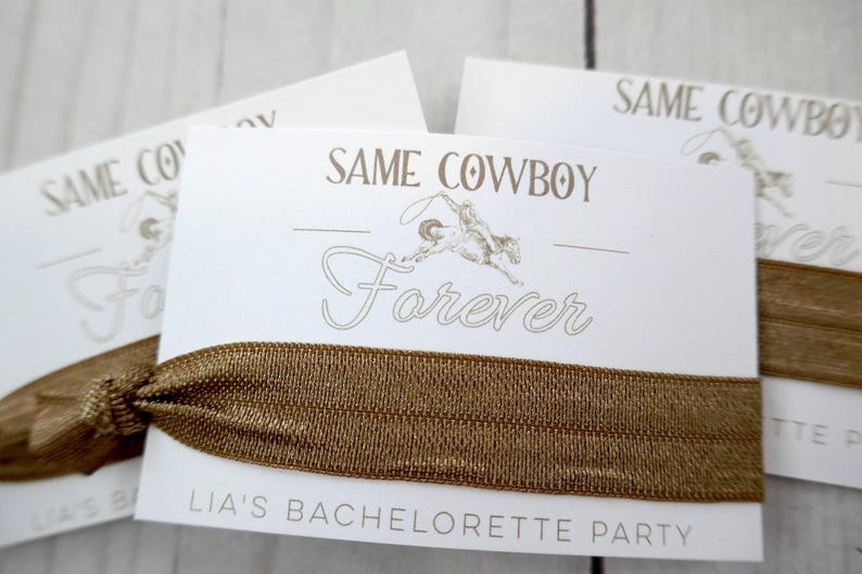K&ouml;nnte beinhalten: Wei&szlig;e Karte mit dem Text "Same Cowboy Forever" und einer kleinen Illustration eines Cowboys, der ein Pferd reitet. Die Karte ist an einem braunen Gummiband befestigt. Der Text "Lia's Bachelorette Party" ist unten auf der Karte gedruckt.