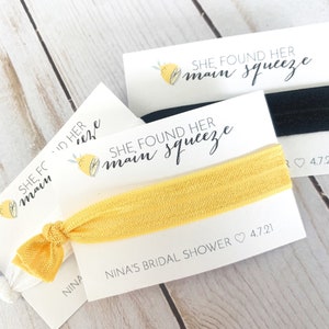 May include: A yellow elastic hair tie with a white card attached. The card has a lemon illustration and the text "SHE FOUND HER MAIN SQUEEZE" and "NINA'S BRIDAL SHOWER 4.7.21".