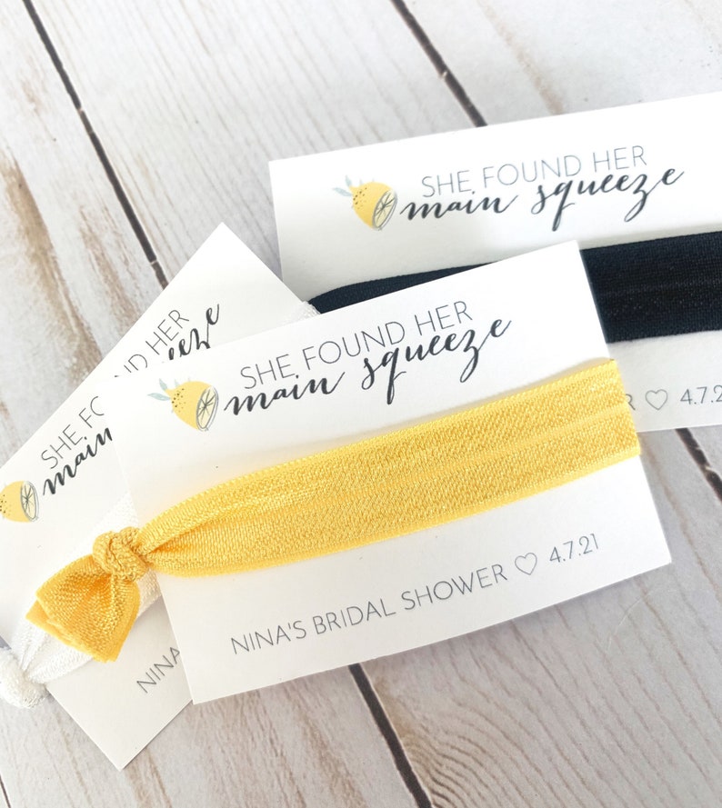 May include: A yellow elastic hair tie with a white card attached. The card has a lemon illustration and the text "SHE FOUND HER MAIN SQUEEZE" and "NINA'S BRIDAL SHOWER 4.7.21".