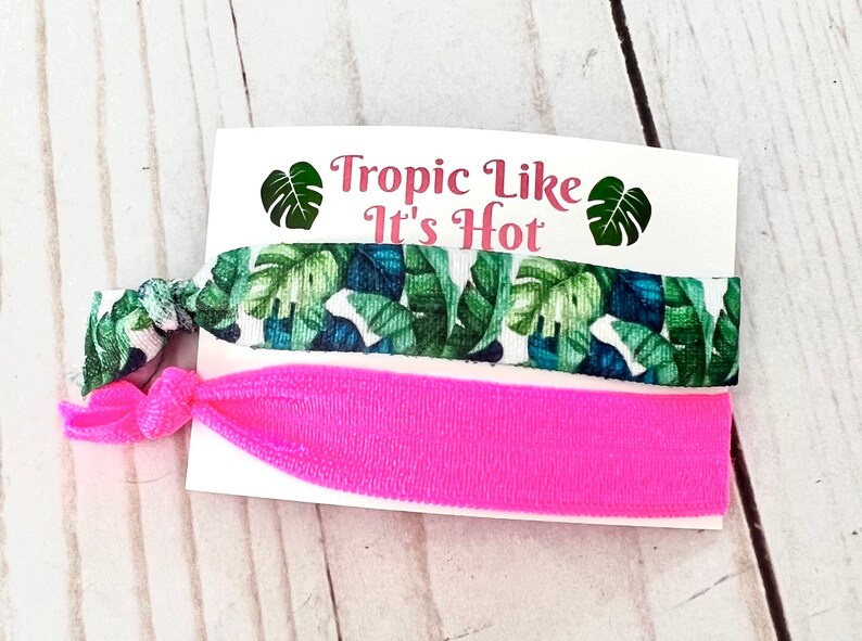 Op de afbeelding: Twee haar elastiekjes, een met een tropische bladerenprint en de andere effen roze. De kaart staat "Tropic Like It's Hot".