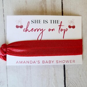 May include: Red elastic hair tie with a white card attached. The card says "She is the cherry on top" and "Amanda's Baby Shower".