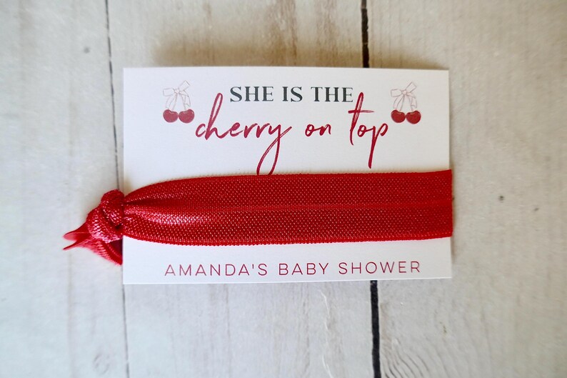 May include: Red elastic hair tie with a white card attached. The card says "She is the cherry on top" and "Amanda's Baby Shower".