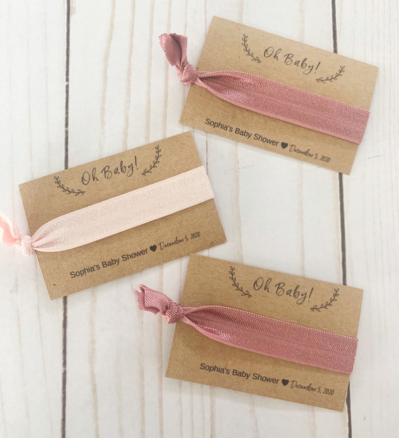 K&ouml;nnte beinhalten: Drei braune Karten-Geschenke mit rosafarbenen Gummib&auml;ndern f&uuml;r Haare. Jede Karte tr&auml;gt den Text "Oh Baby!" und "Sophia's Baby Shower December 5, 2020".