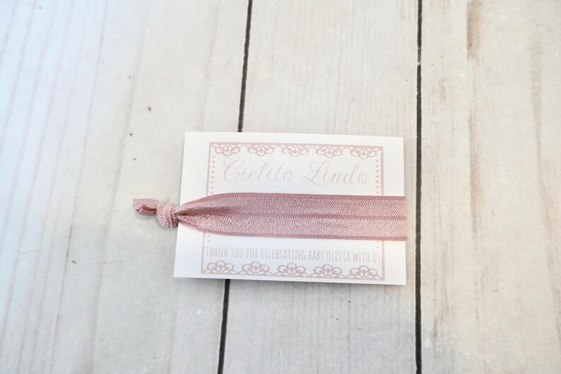K&ouml;nnte beinhalten: Eine wei&szlig;e Karte mit einem rosa Band darum gebunden. Die Karte hat einen dekorativen Rand und den Text "Cielito Lindo" in Kursivschrift. Der Text unter dem Rand lautet "Vielen Dank, dass Sie Baby Olivia mit uns gefeiert haben."