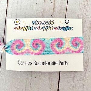 May include: A white card with the text "She Said alright alright alright" and a tie-dye elastic hair tie. The card also says "Cassie's Bachelorette Party".
