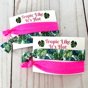 Op de afbeelding: Twee sets haarbandjes met een tropische bladerenprint en een effen roze haarbandje. Elke set is verpakt op een witte kaart met de tekst "Tropic Like It's Hot" en een groene tropische bladerenafbeelding.
