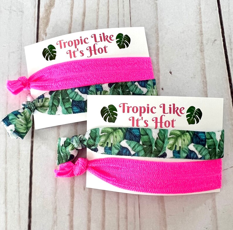 Op de afbeelding: Twee sets haarbandjes met een tropische bladerenprint en een effen roze haarbandje. Elke set is verpakt op een witte kaart met de tekst "Tropic Like It's Hot" en een groene tropische bladerenafbeelding.