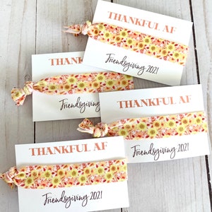 May include: Four white cards with hair ties. Each card has the words "THANKFUL AF" in copper-colored text and "Friendsgiving 2021" in black script. The hair ties have a yellow and orange sunflower pattern.