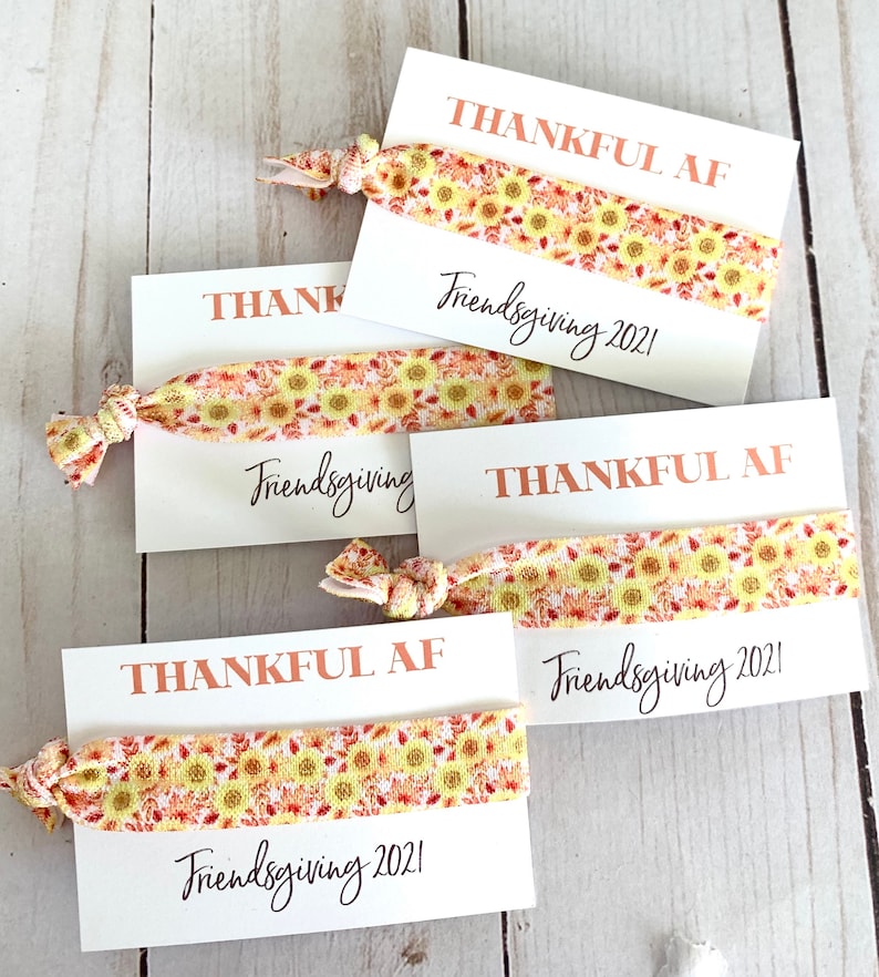 May include: Four white cards with hair ties. Each card has the words "THANKFUL AF" in copper-colored text and "Friendsgiving 2021" in black script. The hair ties have a yellow and orange sunflower pattern.