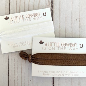 May include: Two white cards with brown text and a brown horseshoe symbol. The text reads "A Little Cowboy U is on the way!" and "Thank you for celebrating Baby Cayden with us!" Each card has a brown elastic hair tie attached.