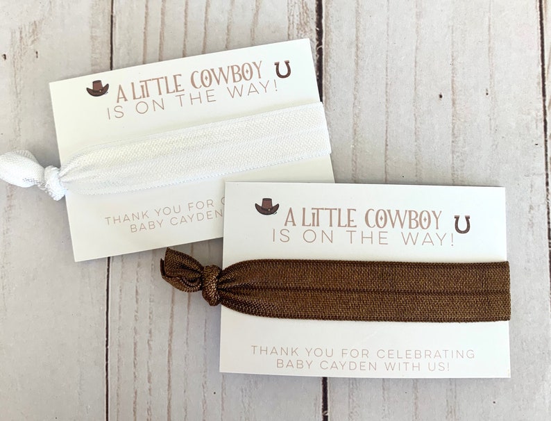 May include: Two white cards with brown text and a brown horseshoe symbol. The text reads "A Little Cowboy U is on the way!" and "Thank you for celebrating Baby Cayden with us!" Each card has a brown elastic hair tie attached.