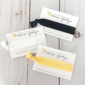 May include: Three white cards with yellow lemon designs and black text that reads "She found her main squeeze" and "Nina's Bridal Shower 4.7.21". Each card has a hair tie attached.