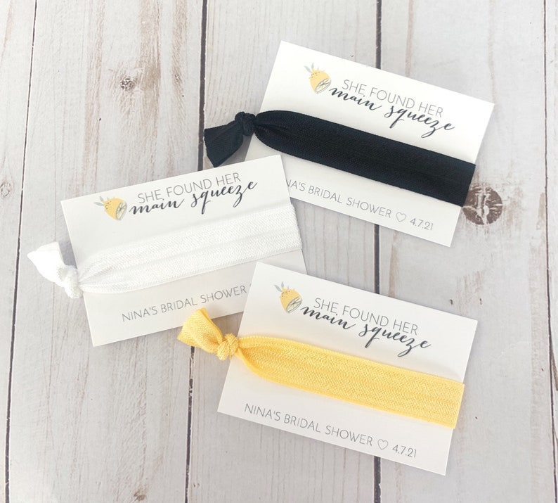 May include: Three white cards with yellow lemon designs and black text that reads "She found her main squeeze" and "Nina's Bridal Shower 4.7.21". Each card has a hair tie attached.
