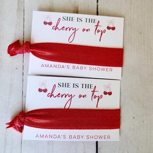 May include: Two white cards with red cherry graphics and the text "She is the cherry on top" and "Amanda's Baby Shower". Each card has a red elastic hair tie attached.