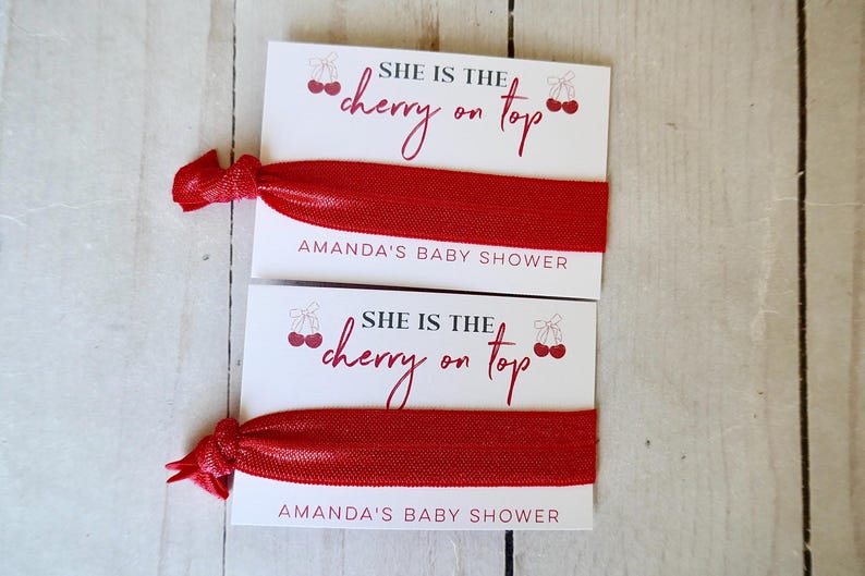 May include: Two white cards with red cherry graphics and the text "She is the cherry on top" and "Amanda's Baby Shower". Each card has a red elastic hair tie attached.