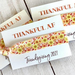 May include: White cards with the text "THANKFUL AF" in orange lettering. Each card has a sunflower patterned hair tie. The words "Friendsgiving 2021" are written in cursive on the cards.