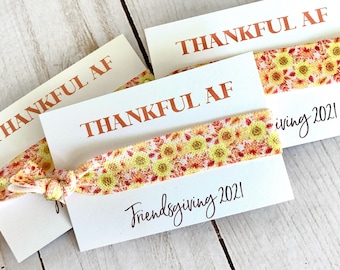 May include: White cards with the text "THANKFUL AF" in orange lettering. Each card has a sunflower patterned hair tie. The words "Friendsgiving 2021" are written in cursive on the cards.