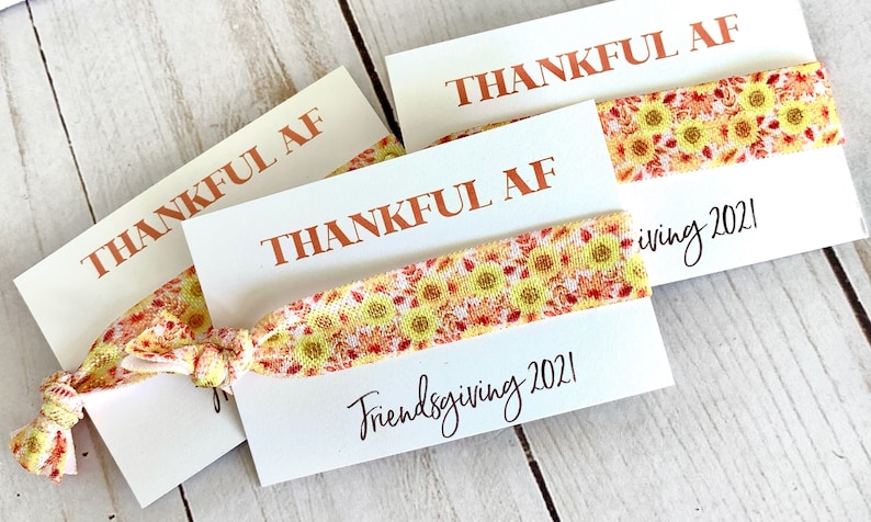 May include: White cards with the text "THANKFUL AF" in orange lettering. Each card has a sunflower patterned hair tie. The words "Friendsgiving 2021" are written in cursive on the cards.