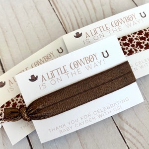 May include: A white card with brown text that reads "A Little Cowboy is on the Way!" with a brown cowboy hat and horseshoe graphic. A brown elastic hair tie is wrapped around the card. The card is on a wooden surface.