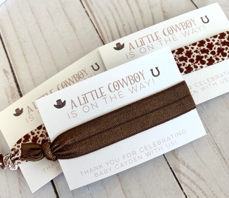 May include: A white card with brown text that reads "A Little Cowboy is on the Way!" with a brown cowboy hat and horseshoe graphic. A brown elastic hair tie is wrapped around the card. The card is on a wooden surface.