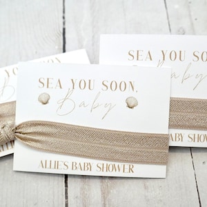 May include: Cards with elastic hair ties for a baby shower. The cards are white with the text "Sea You Soon, Baby" and a seashell graphic. The hair ties are a light brown color. The text "Allie's Baby Shower" is also printed on the cards.