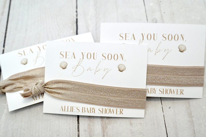 May include: Cards with elastic hair ties for a baby shower. The cards are white with the text "Sea You Soon, Baby" and a seashell graphic. The hair ties are a light brown color. The text "Allie's Baby Shower" is also printed on the cards.