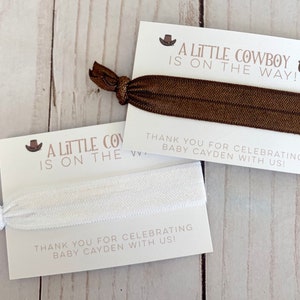 May include: Two white cards with brown text and a brown and white elastic hair tie. The text on the cards reads "A Little Cowboy is on the Way!" and "Thank you for celebrating Baby Cayden with us!"