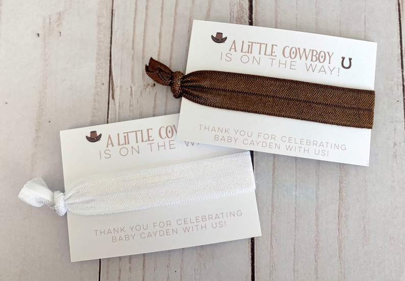 May include: Two white cards with brown text and a brown and white elastic hair tie. The text on the cards reads "A Little Cowboy is on the Way!" and "Thank you for celebrating Baby Cayden with us!"