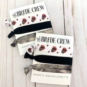K&ouml;nnte beinhalten: Set aus drei Haargummis mit wei&szlig;em Hintergrund und einem Muster aus roten Lippen und einem Rock'n'Roll-Handsymbol. Die Haargummis sind mit einer Karte verpackt, auf der "Bride Crew" und "Anne's Bachelorette" steht.