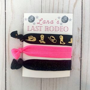 May include: A set of three hair ties. One black hair tie with gold cowboy boot and hat designs, one bright pink hair tie, and one black glitter hair tie. The card reads "Lana's LAST RODEO".