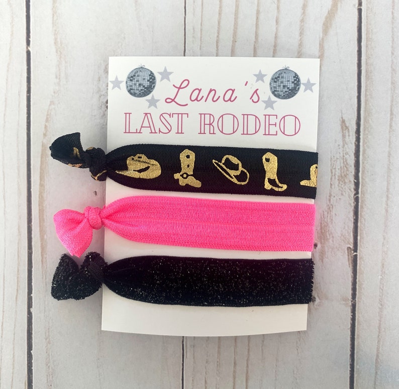 May include: A set of three hair ties. One black hair tie with gold cowboy boot and hat designs, one bright pink hair tie, and one black glitter hair tie. The card reads "Lana's LAST RODEO".