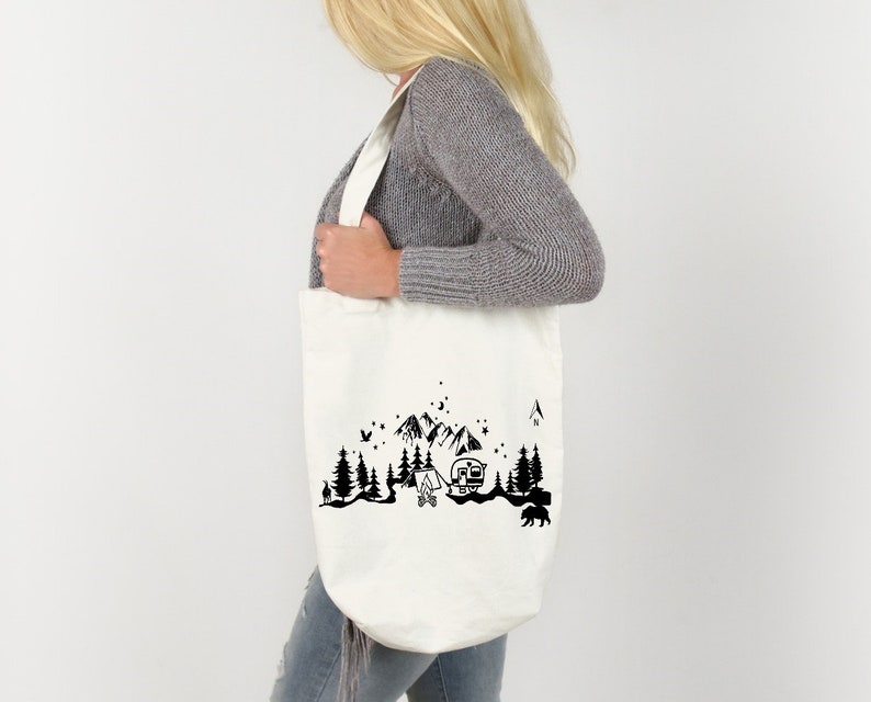 Puede incluir: Una bolsa de lona blanca con un dise&ntilde;o de silueta negra de una caravana, monta&ntilde;as, &aacute;rboles y una fogata. El dise&ntilde;o recuerda una escena de campamento.