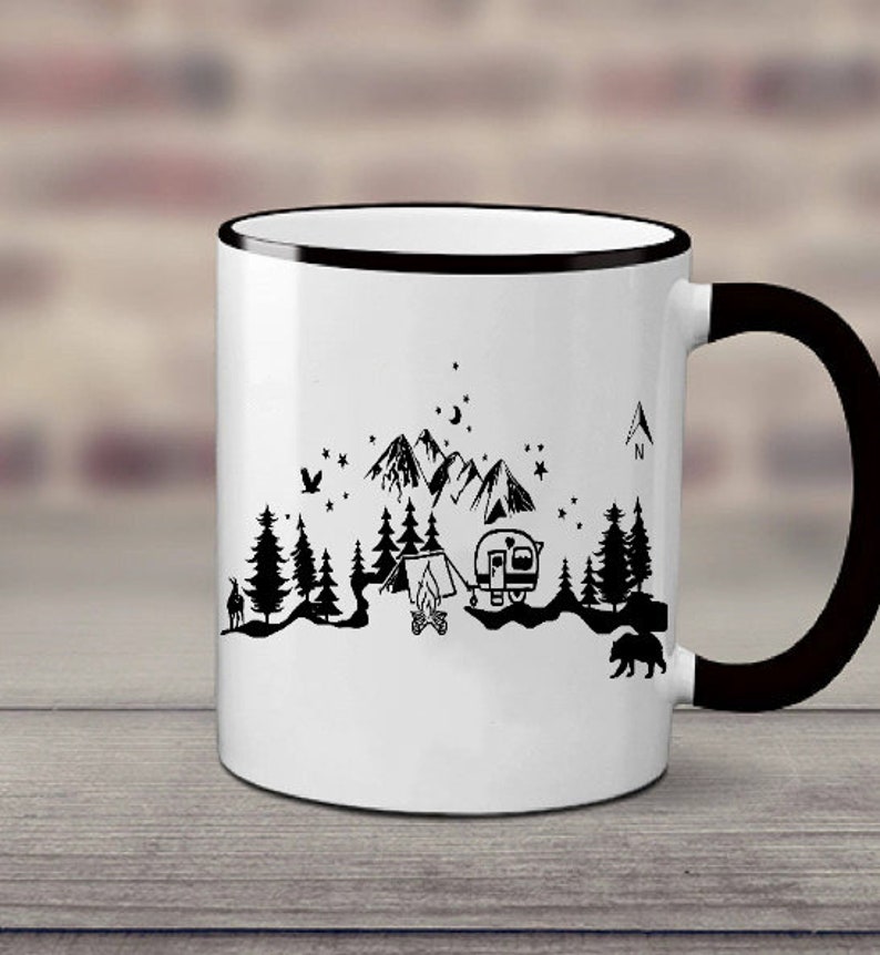 Puede incluir: Taza de cer&aacute;mica blanca con borde y asa negros. La taza presenta una ilustraci&oacute;n en blanco y negro de una escena de campamento con monta&ntilde;as, &aacute;rboles, una tienda de campa&ntilde;a, una caravana y una fogata. El texto "N" es visible en la esquina superior derecha de la taza.