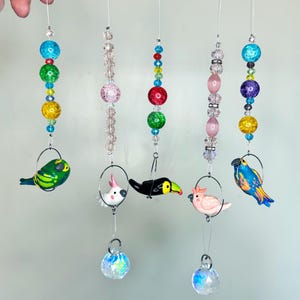 May include: A set of six colorful beaded suncatchers with glass bird and animal charms. Each suncatcher features a crystal ball at the bottom. The charms include a toucan, a parrot, a cockatoo, and two small birds.