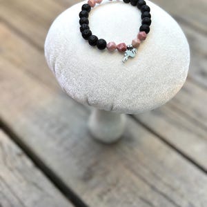 Peut inclure: Un bracelet de perles composé de perles de lave noire, de pierres précieuses roses et d'un charm argenté. Le bracelet est présenté sur un piédestal blanc en forme de champignon, sur fond de bois. Le charm est un petit oiseau stylisé.