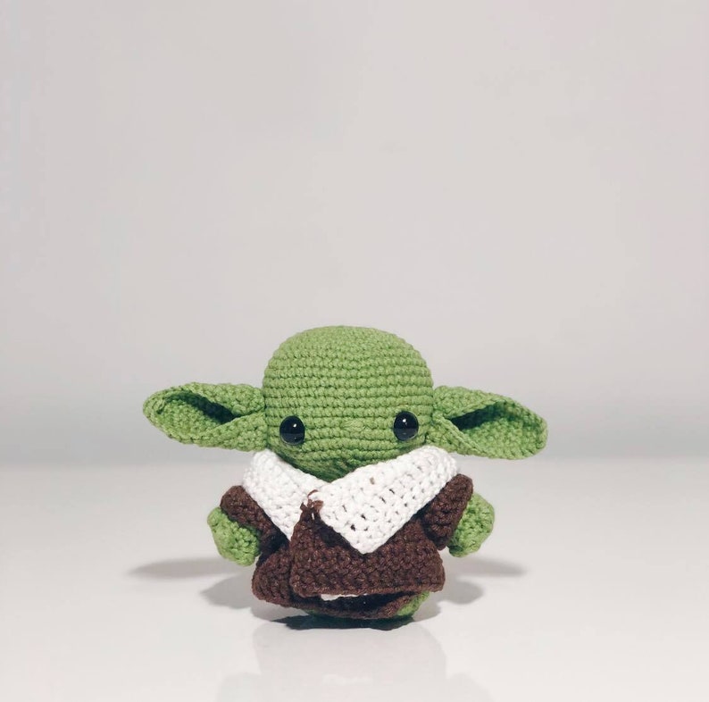 May include: A green crocheted plush toy with large ears and a brown and white crocheted coat. The toy has black eyes and a small mouth.