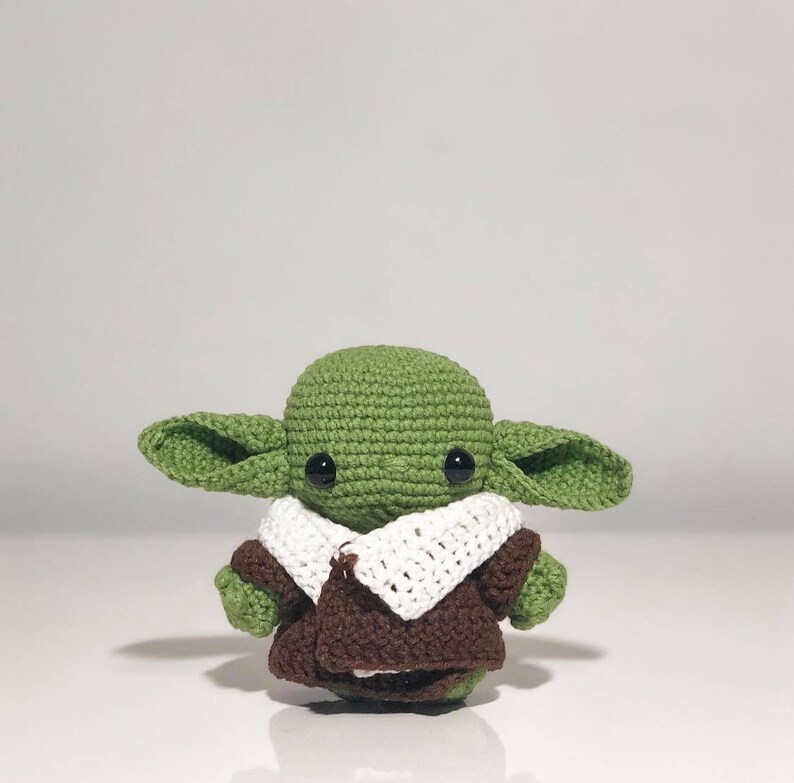 May include: A green crocheted plush toy with large ears and big eyes. The toy is wearing a brown and white crocheted jacket.