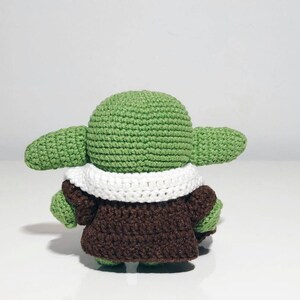 May include: A green crocheted plush toy with a white crocheted collar and a brown crocheted coat. The toy is standing on a white surface.