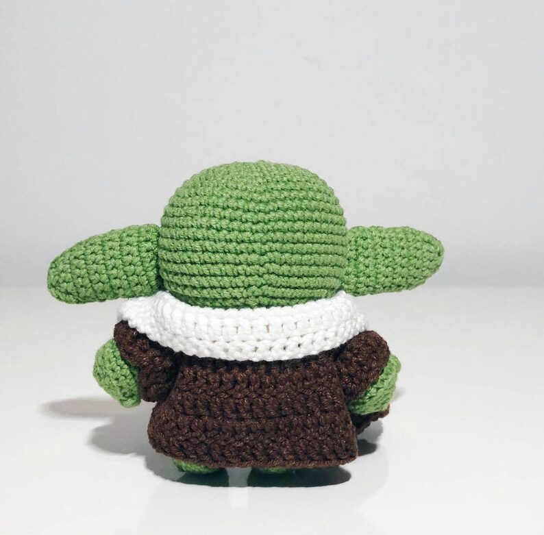 May include: A green crocheted plush toy with a white crocheted collar and a brown crocheted coat. The toy is standing on a white surface.