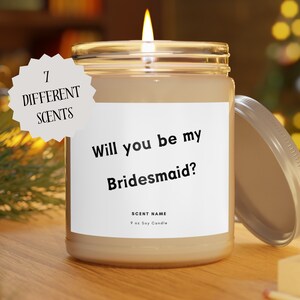 May include: A lit soy candle in a glass jar with a silver lid. The label reads "Will you be my Bridesmaid?" and "7 Different Scents". The candle is 9 oz.