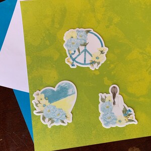 May include: Three stickers with a peace sign, a heart, and a woman with flowers. The peace sign is surrounded by yellow and blue flowers. The heart is blue and yellow with flowers on the side. The woman is wearing a white dress and has brown hair. All three stickers have a white background.