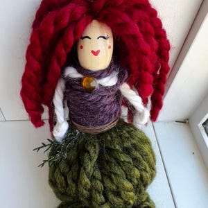 May include: A handmade yarn doll with a wooden head, red yarn hair, a purple yarn bodice, a white yarn scarf, a brown yarn belt, and a green yarn skirt.