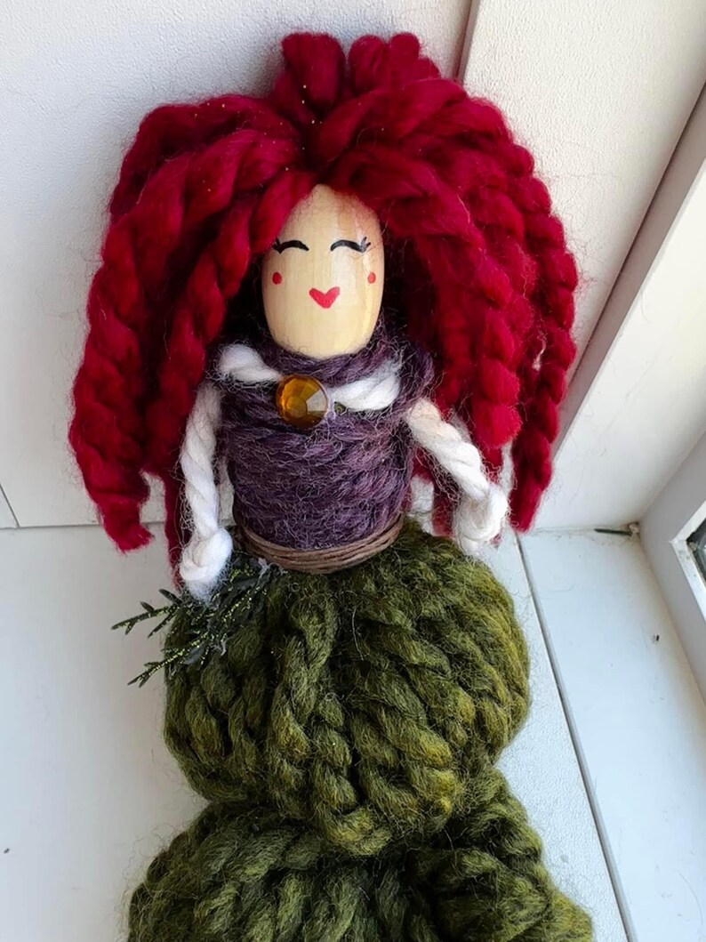 May include: A handmade yarn doll with a wooden head, red yarn hair, a purple yarn bodice, a white yarn scarf, a brown yarn belt, and a green yarn skirt.