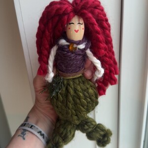 May include: A handmade yarn doll with red yarn hair, a purple yarn bodice, a brown yarn belt, and a green yarn tail. The doll has a painted face with red cheeks and a smiling mouth.