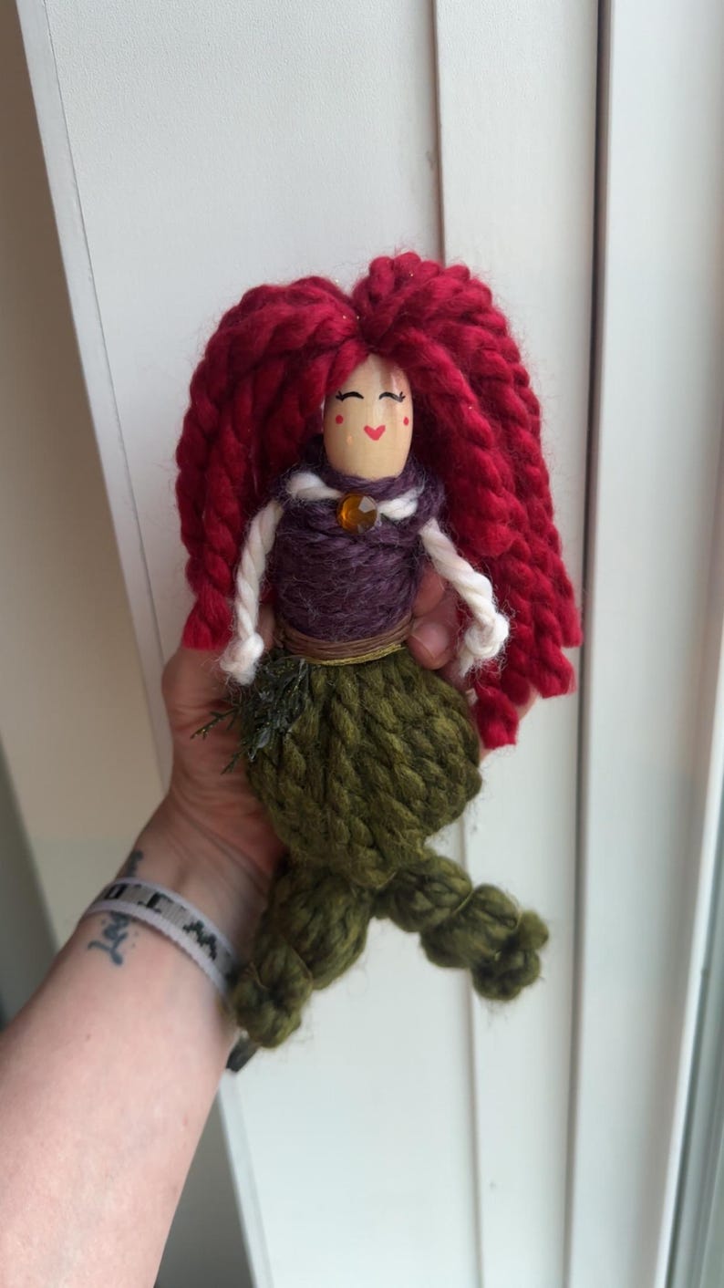 May include: A handmade yarn doll with red yarn hair, a purple yarn bodice, a brown yarn belt, and a green yarn tail. The doll has a painted face with red cheeks and a smiling mouth.