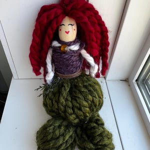 May include: A handmade yarn doll with red yarn hair, a purple yarn bodice, and a green yarn tail. The doll has a painted face with red cheeks and a brown bead for a nose.