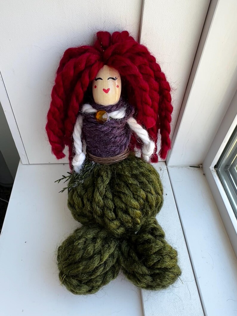 May include: A handmade yarn doll with red yarn hair, a purple yarn bodice, and a green yarn tail. The doll has a painted face with red cheeks and a brown bead for a nose.