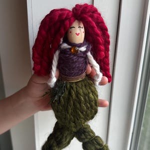 May include: A handmade yarn doll with red hair, a purple bodice, and a green mermaid tail. The doll has a painted face with red cheeks and a smile.